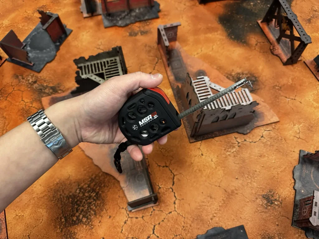 flgtapemeasure2 Frontline Gaming Person holding a small retractable tape measure above a tabletop wargame terrain with orange cracked ground and ruined buildings measuring distance