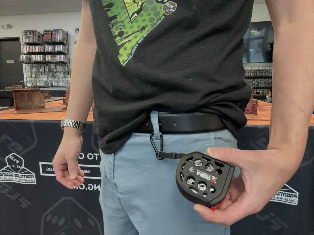 flgtapemeasure3 Frontline Gaming Man in a black Tshirt shows a black circular dice holder with multiple holes clipped to his belt