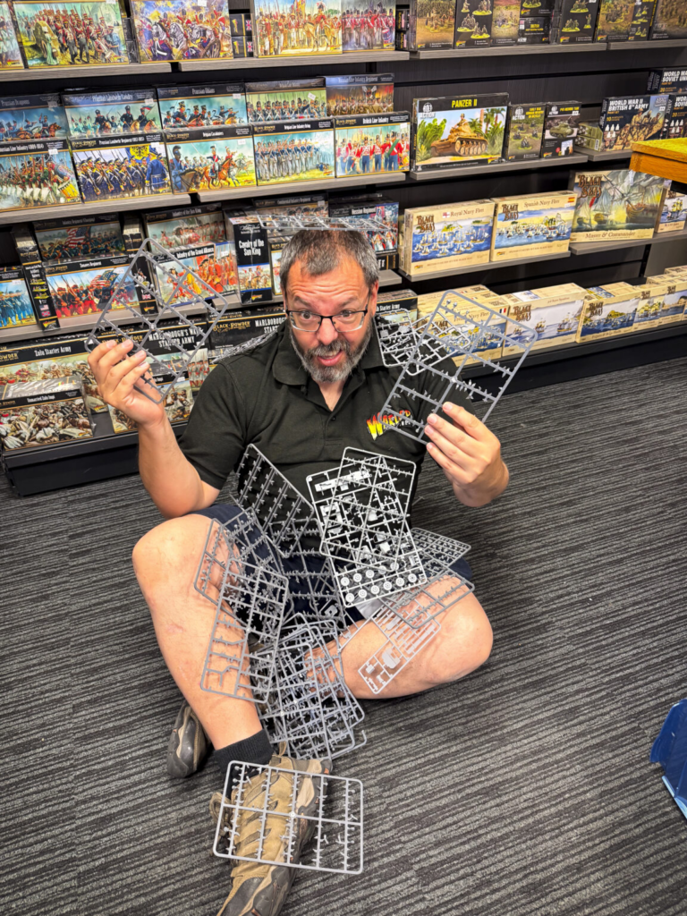 image Frontline Gaming Man sits on the floor in a hobby store holding multiple gray plastic model sprues with boxed miniatures on the shelves behind him