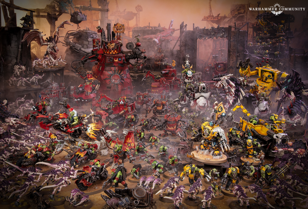 Extensive Warhammer diorama battle in a ruined city with red and yellow armored walkers and hundreds of miniatures amid smoke Warhammer Community logo top right