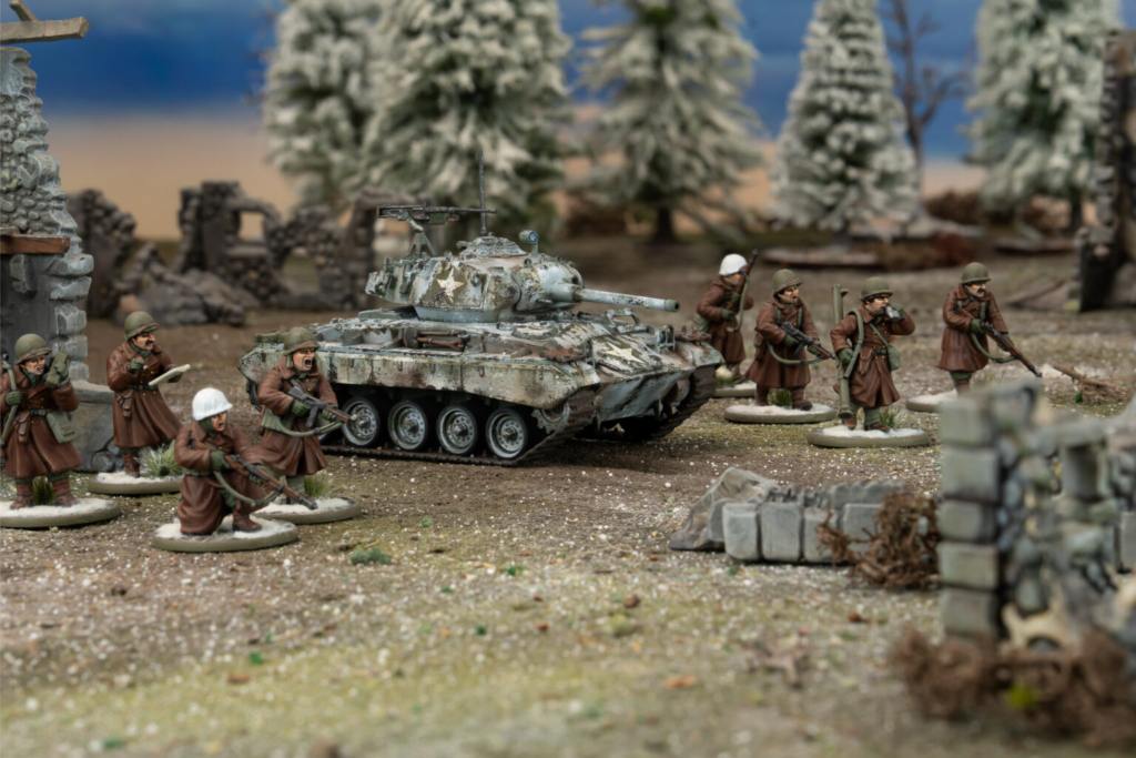 Scale model diorama of WW2 soldiers advancing around a weathered tank in a ruined village with snow covered trees in the background
