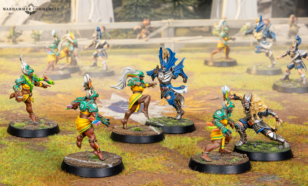 Top left Warhammer Community logo over a chaotic tabletop battle of painted miniatures facing off on a grassy base with a central blue armored figure confronting a green yellow warrior