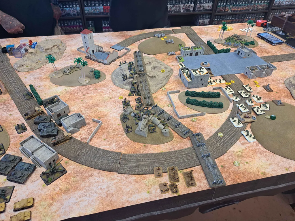Tabletop wargame battlefield with model tanks trenches ruins and a circular railway track on a desert toned gaming table miniature soldiers maneuvering across terrain