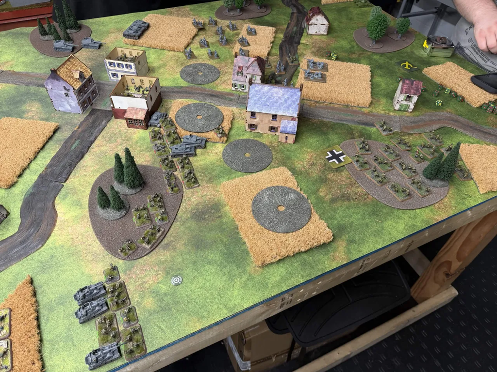 Tabletop wargame scene with miniature tanks infantry and ruined buildings on a grassy battlefield with fields and roads