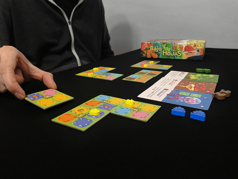 image Frontline Gaming A persons hand places a colorful game tile on a board setup with several tile cards and small yellow pieces on a black table and the game box in the background
