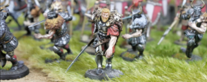 Close up of a fantasy warrior miniature in armor with a red cape and raised sword leading a group on a grassy battlefield diorama