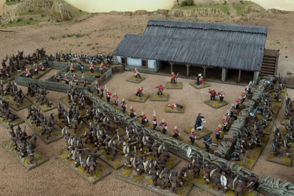 Miniature wargame battlefield with red coated troops defending a stone wall and thatched building amid brown uniform forces on a dusty terrain