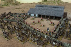 Miniature wargame battlefield with red coated troops defending a stone wall and thatched building amid brown uniform forces on a dusty terrain