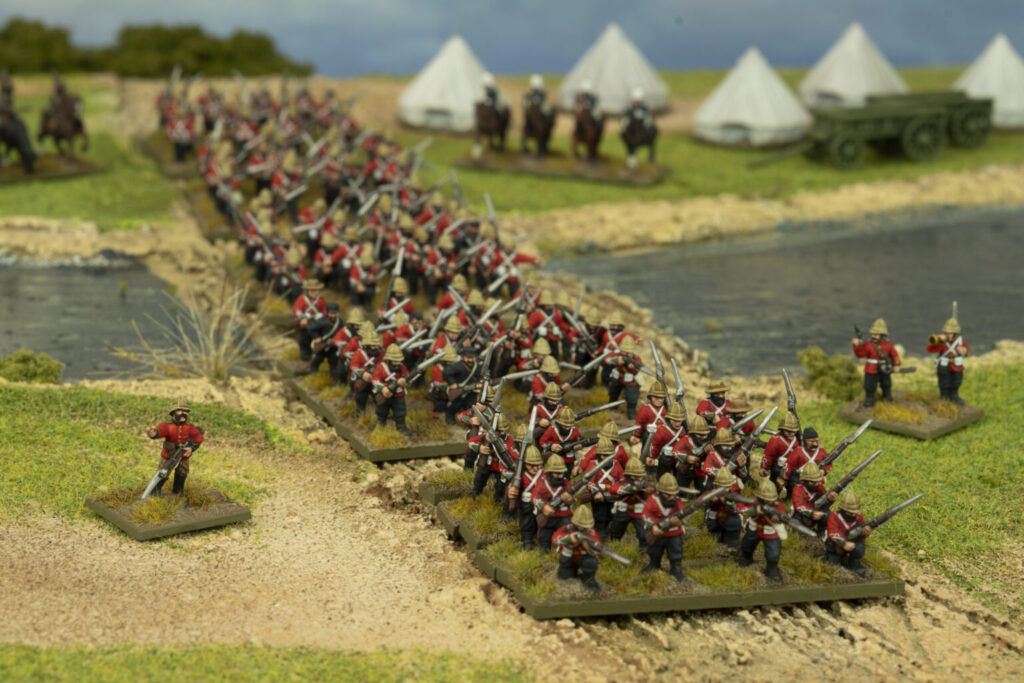 Miniature soldiers in red coats marching in formation across a dirt path by a river with tents and a wagon in the background as part of a diorama battle scene