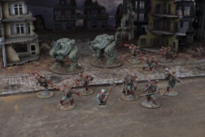 Tabletop wargame scene on a ruined city street with two large teal armored walkers facing groups of red clad orc like miniatures and civilians