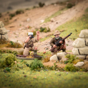 Two toy soldiers in a diorama one kneels with binoculars raised the other crouches behind rocks with a rifle in a desert like landscape