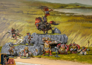 Tabletop fantasy battle scene with vividly painted miniatures a giant armored rider on a rock pedestal faces a line of orc soldiers on a grassy plain