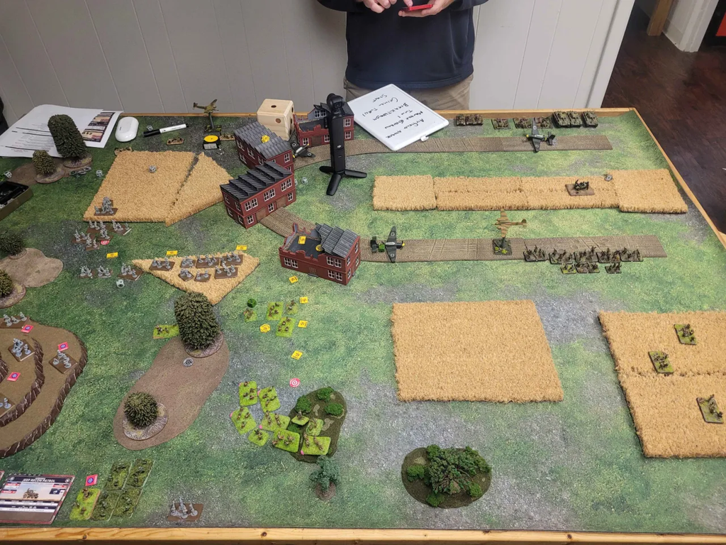 Tabletop wargame setup with miniatures buildings and fields arranged on a model battlefield plus a whiteboard and player notes nearby