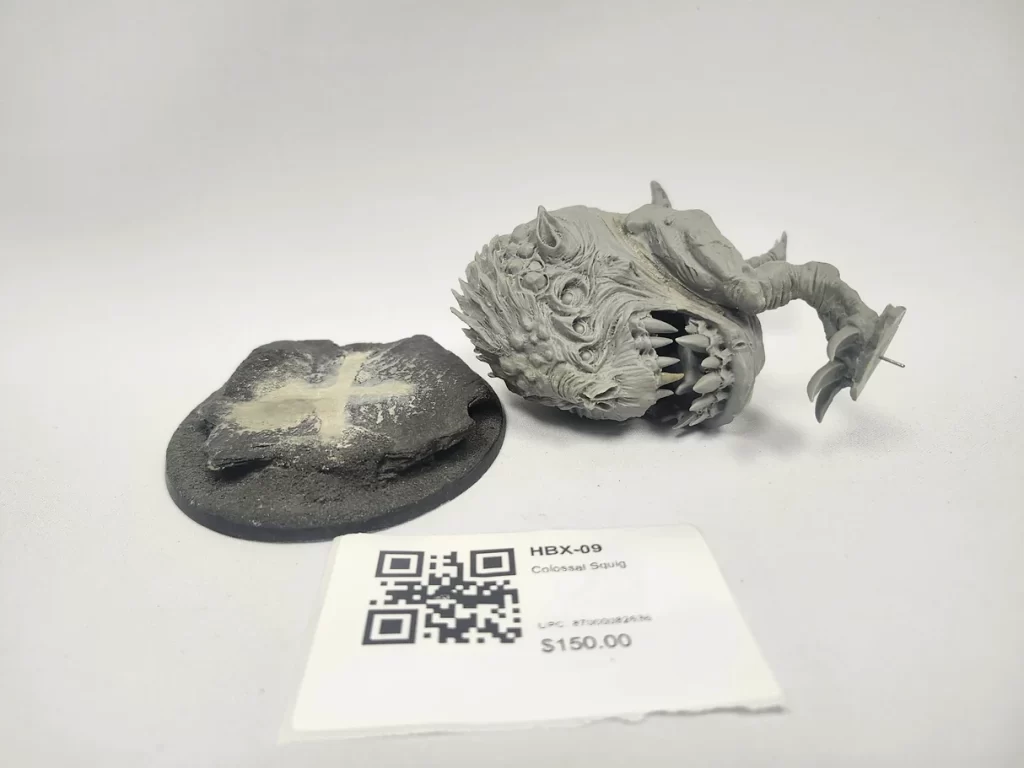 Gray resin miniature of a monstrous squid headed creature with jagged teeth lying beside a round base on a white backdrop